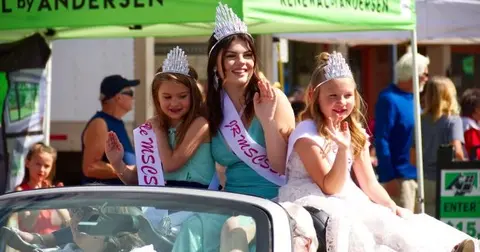 Three people wearing tiaras and sashes wave from a convertible in a parade.
