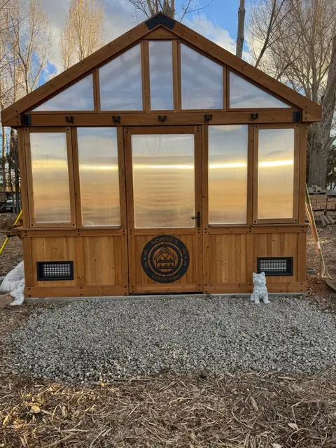 A wooden greenhouse stands outdoors on a gravel pad.