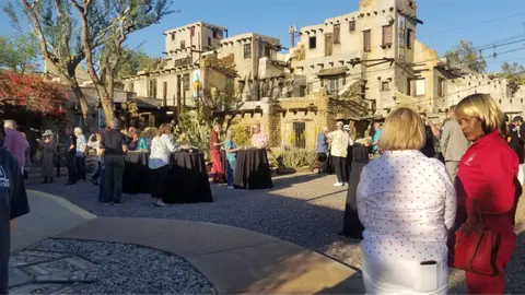 People gather in a courtyard outside pueblo-style buildings.