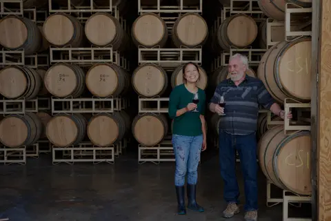 Two people holding wine glasses stand in front of stacks of wine barrels.