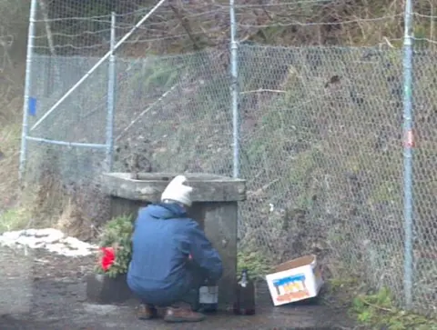A person crouching near a concrete structure against a chain-link fence.