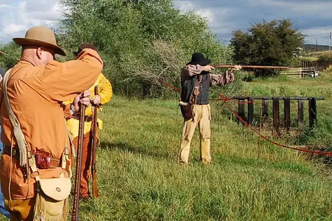 People in period costume participate in a shooting activity outdoors.