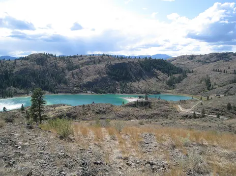 A scenic view of a turquoise lake nestled among dry hills and pine trees.