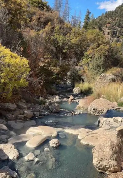 Natural hot springs cascade down a rocky mountain slope surrounded by autumn trees.