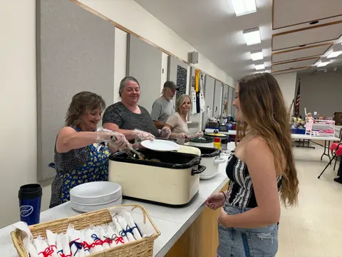 Volunteers serve food from roaster ovens to a young woman indoors.