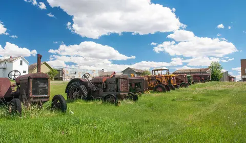 A line of old, rusty tractors sits in a grassy field in front of rural buildings.