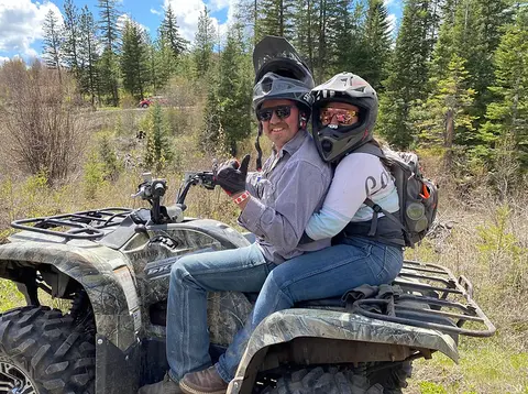 A couple riding an ATV in a wooded area.