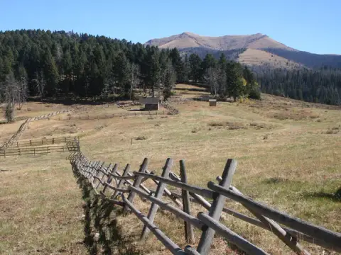 A wooden fence leads across a grassy meadow toward small cabins with mountains and a forest in the background.