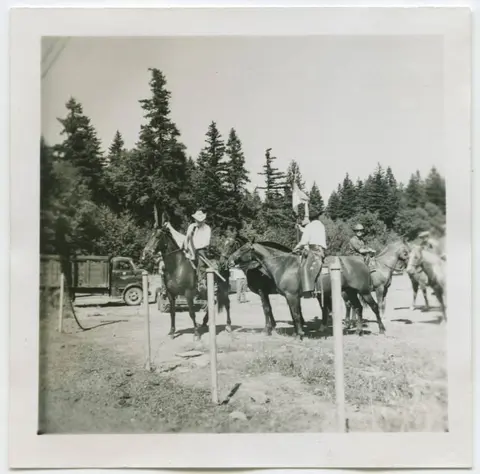 A historic black and white photograph showing people on horseback outdoors near trees and a truck.