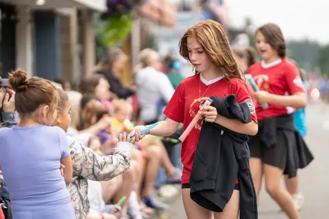 A young girl in a red shirt hands out treats to children along a street.