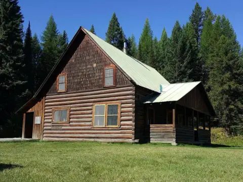 A rustic log cabin stands in a grassy clearing surrounded by tall evergreen trees under a clear blue sky.