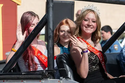 Three young women wearing tiaras and sashes wave from a vehicle during a parade.