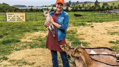 A woman holds a small goat in a pasture with several other goats around her.