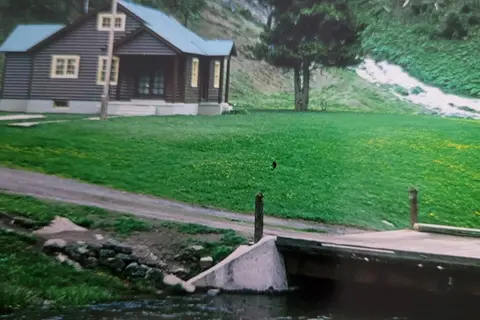 A dark wooden building sits on a lawn beside a stream and a small bridge.