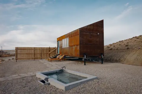 A small rust-clad tiny home sits on a gravel lot next to a small concrete pool in an arid landscape.