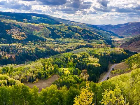 An aerial view of a mountain valley with autumn-colored forests and a winding road.