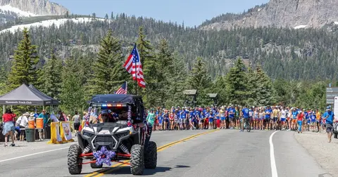 An off-road vehicle decorated with American flags leads a crowd of people gathered on a paved road.