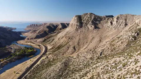 An aerial landscape shows a winding river, railroad tracks, and arid mountains under a blue sky.