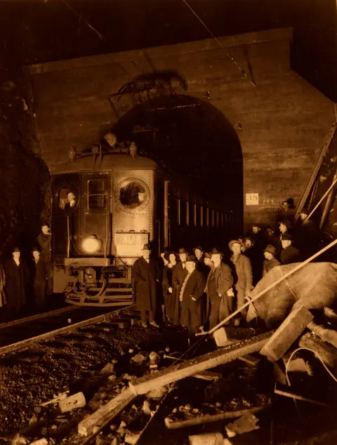 An old sepia photograph showing a train emerging from a tunnel with people standing nearby.