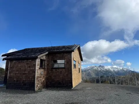 A small wooden cabin stands outdoors with a view of snowy mountains.