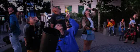 People look up at the sky near telescopes outdoors.