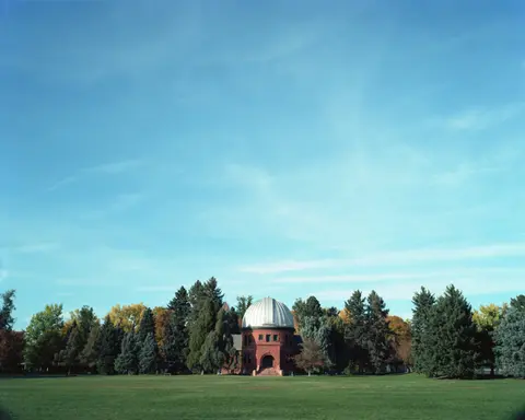 A red brick observatory building with a dome stands behind a large grassy lawn and trees.