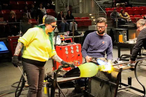 People perform glassblowing with torches inside a workshop.