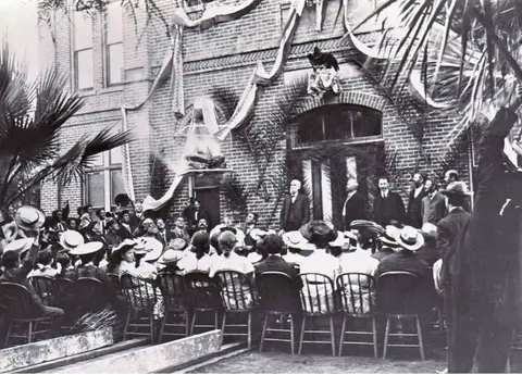 A historic black and white photograph showing a crowd of people gathered outside a decorated building.