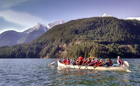 A group of people in a large canoe paddle on a lake with mountains in the background.