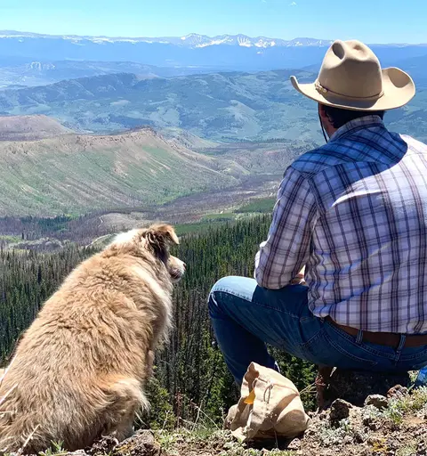 A person in a cowboy hat and plaid shirt sits with a dog looking out over a mountain landscape.