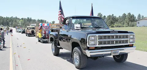 A classic black pickup truck leads a parade down a street with spectators watching.