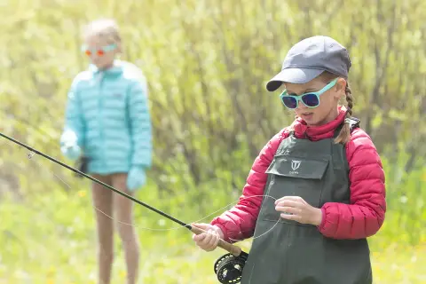 A young girl holding a fishing rod outdoors with another child in the background.