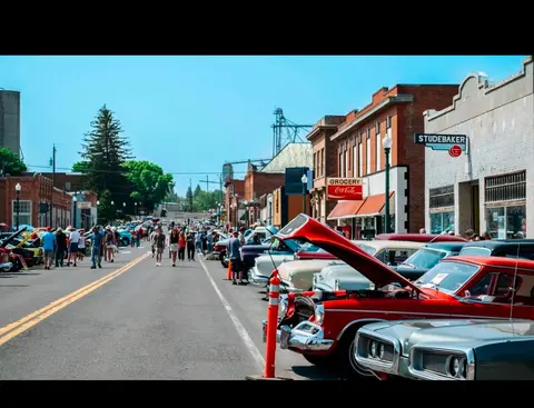 Classic cars are parked along a street lined with buildings while people walk by.