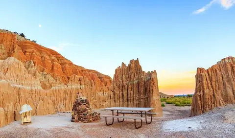 A picnic table and trash can in a desert landscape with dramatic eroded rock formations.