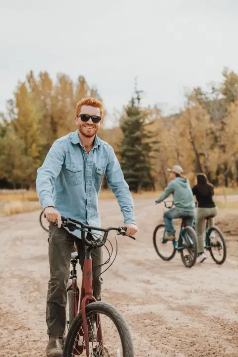 A smiling man rides a bicycle on an outdoor dirt path with other people riding behind him.
