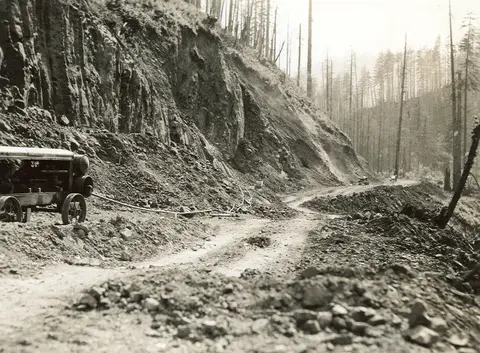A historic black and white photograph showing a dirt road under construction alongside a rocky cliff and forested hillside, with machinery in the foreground.