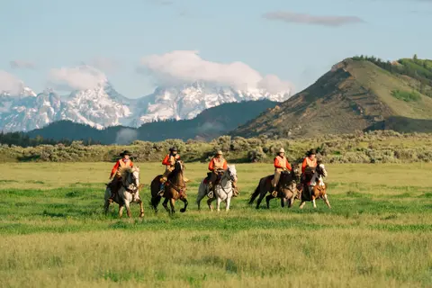Five people ride horses across a grassy field with mountains in the background.