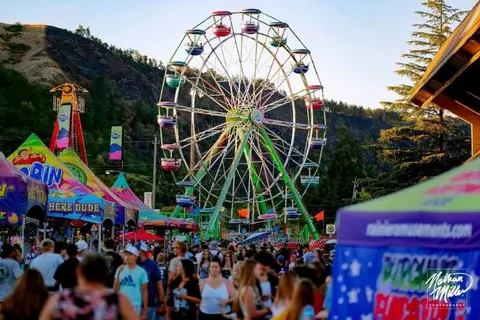 A large Ferris wheel stands above a crowded carnival with colorful tents and people walking around.