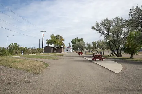 An outdoor park with picnic tables, RV parking spots, and buildings in the distance under a partly cloudy sky.