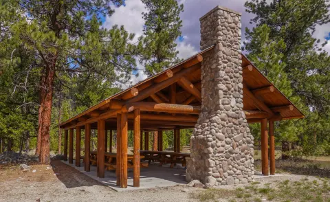 A rustic wooden pavilion with a large stone fireplace stands in a forest.