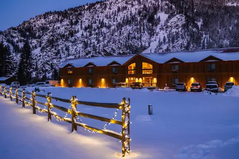 A wooden lodge at dusk with snow on the ground and a lit fence in the foreground.