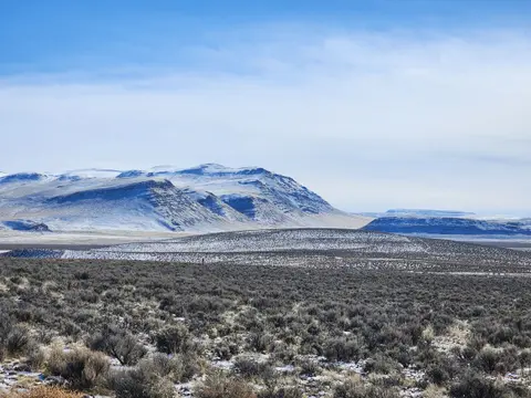 A landscape showing snow-covered mountains and desert scrubland under a blue sky.
