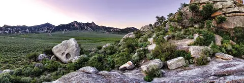 A wide landscape photograph showing rocky terrain, green valleys, and distant mountains at dusk.