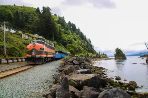 A train travels along a coastal track next to a rocky shoreline and forested hillside.