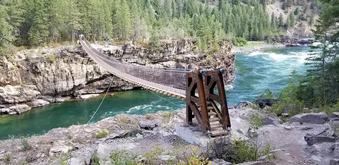 A suspension bridge spans a river flowing between rocky banks and forests.
