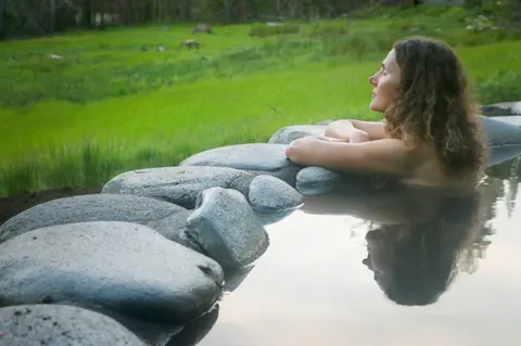 A woman relaxes in an outdoor hot spring pool surrounded by stones.