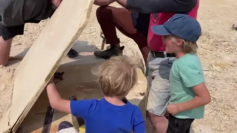 People lifting a large stone slab outdoors while children watch.