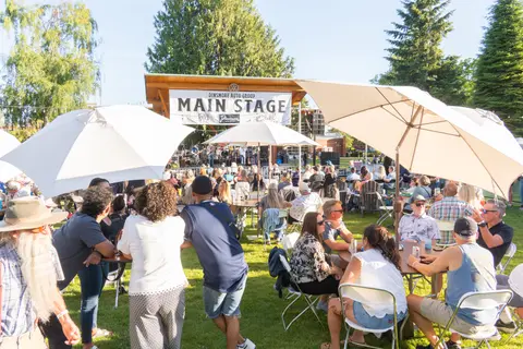 An outdoor concert venue with people seated at tables under umbrellas watching a stage.