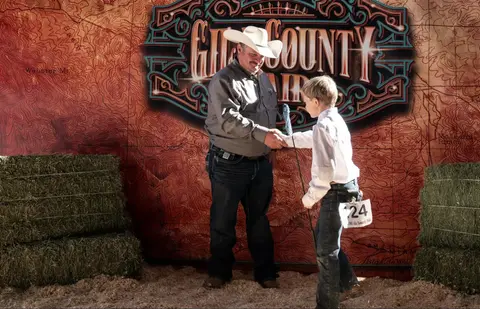 A man wearing a cowboy hat shakes hands with a boy holding a show stick in front of a fair backdrop.