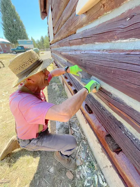 A man applies chinking between the logs of a wooden cabin exterior.